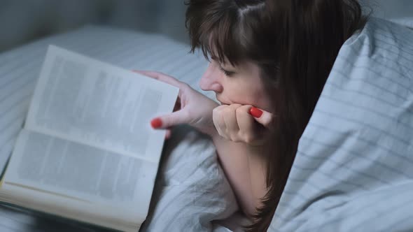 Brunette Woman Covered Herself with a Blanket Lies on the Bed Near the Window and Reads a Book at