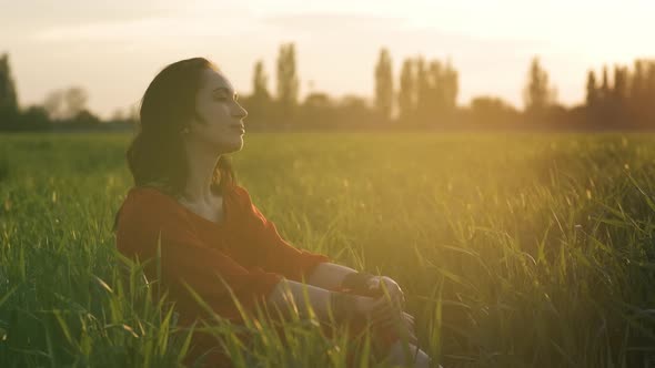 Portrait of a Beautiful Young Hippie Woman in a Red Dress Sits in the Grass of Young Wheat at Sunset alt