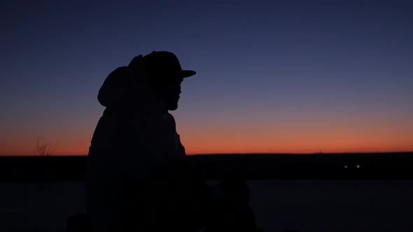 A young man in a cap sits thoughtfully at sunset and dreams alt