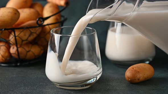 Pouring vegan potato milk in glass and potato in bowl on dark background. Vegan milk.