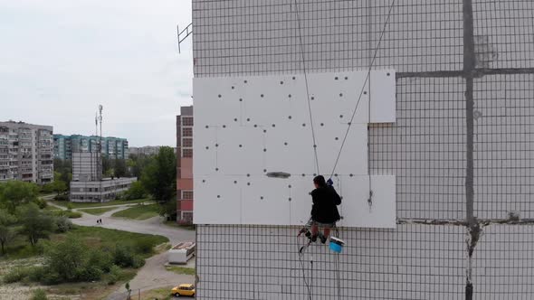 Aerial View on Industrial Climber Suspended on Ropes Performs Work on Insulation of Facade of High alt
