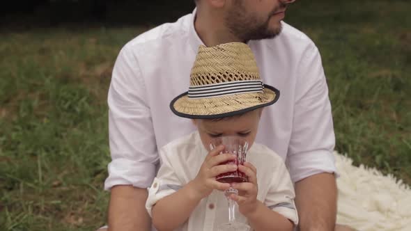 A Young Child Drinks Juice From a Glass at a Picnic with His Parents in the Park