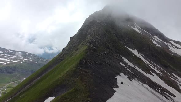 Drone shot (rotating) of a Dark Mountain, partially covered in snow and in the Clouds alt