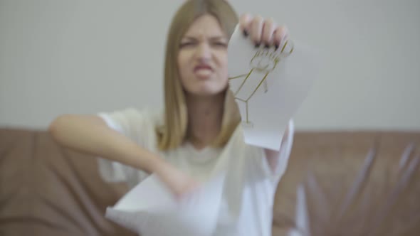 Portrait of Sad Young Woman Tearing Apart Children's Drawing with the Image of Mom and Dad alt