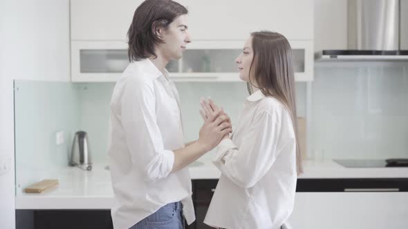 Side View of Happy Young Lovers Holding Hands and Kissing in Kitchen at Home. Portrait of Smiling alt