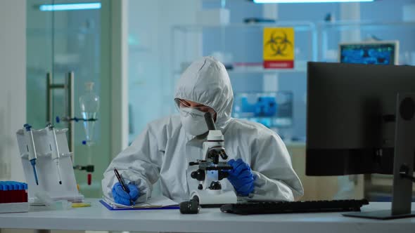Biologist in Ppe Suit Working on Microscope and Writing Information on Clipboard alt