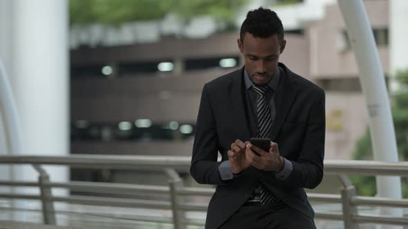 Young African Businessman Thinking While Using Phone Outdoors alt