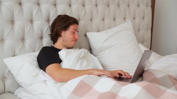 Young Guy Lying in the Bed in the Morning, Yawn and Start Working on Notebook alt