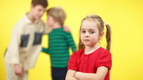 Sad Charming Little Girl with Crossed Hands Looking at Camera As Blurred Boys Mocking at Yellow alt
