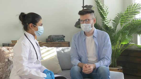 Young female doctor in face mask is talking to male patient at home while man is couching alt