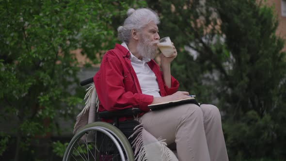 Handicapped Old Caucasian Bearded Man Drinking Cappuccino Writing in Diary with Pen Outdoors alt