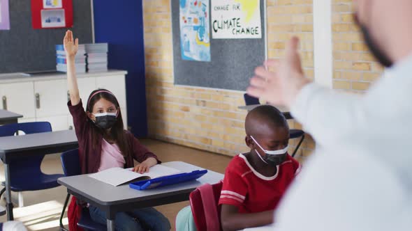 Diverse schoolchildren sitting in classroom raising hands during lesson, all wearing face masks alt