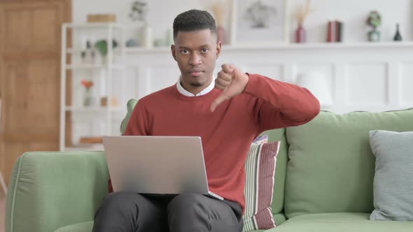 African Man Showing Thumbs Down Sign While using Laptop on Sofa alt