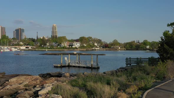A low angle drone shot over a quiet park, heading towards calm waters with New Rochelle in the backg alt
