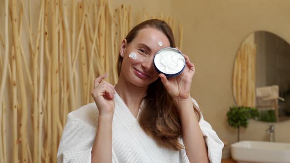 Smiling Beautiful Woman in a Bathrobe Puts a Skin Care Cream on Her Face alt