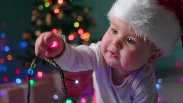 Portrait of a funny happy baby in a Christmas hat, chewing festive lights. Slow motion. alt