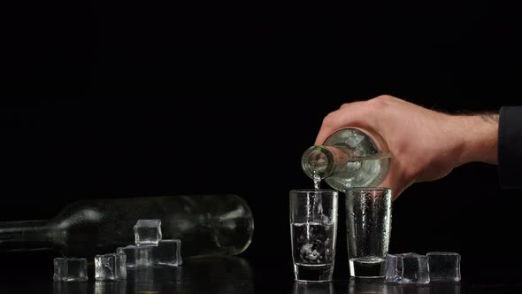 Bartender Pouring Up Vodka From Bottle Into Two Shots Glasses with Ice Cubes on Black Background alt