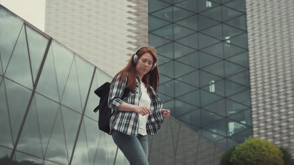 A Cheerful Caucasian Redhaired Girl Dances in Headphones with a Briefcase Against Office Buildings