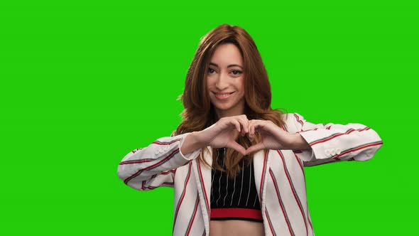Pretty young woman poses on green screen showing heart gesture and blowing kiss alt