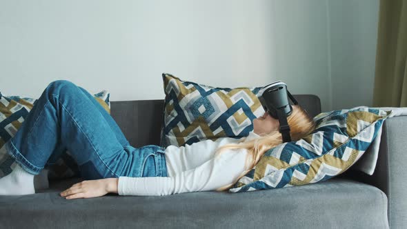 Young Blonde European Girl Lying on the Sofa Wearing Virtual Reality Headset Playing a Game alt