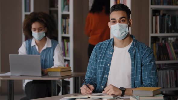 Students Sitting in Library Studying Young Happy Guy in Medical Mask Sits at Desk Preparing for Exam alt