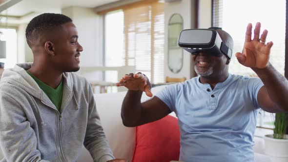 African american senior father sitting on a couch using a vr headset with his teenage son smiling alt