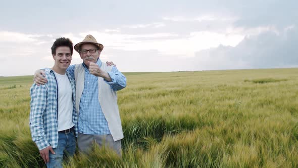 Happy Senior Farmer Embraces Young Son and They Smile at Camera in Barley Field alt