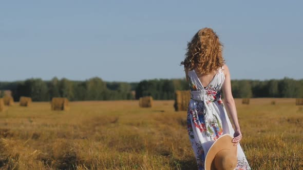 Beautiful Redhaired Woman in a White Dress and Hat Walks Away on the Background of the Field alt