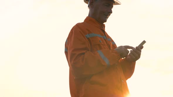 Surveyor in Orange Jumpsuit and Helmet Types on Smartphone alt