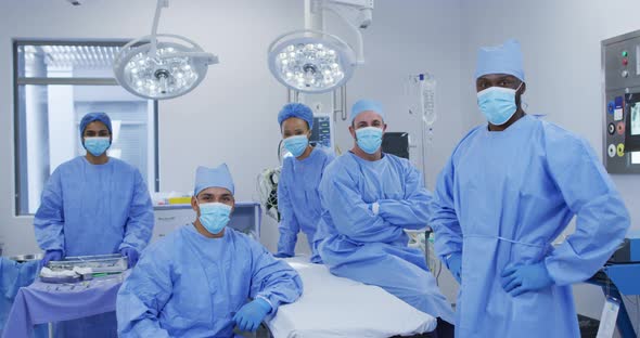 Diverse male and female doctors wearing face masks standing in operating theatre smiling to camera alt