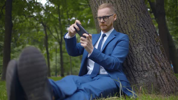 Young Businessman Playing Game on Smartphone Relaxing Under Tree in Park