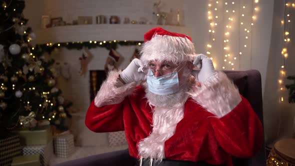 Funny Santa Claus Puts on a Protective Medical Mask While Sitting in a Chair By the Fireplace and