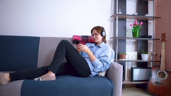 A woman lying on the couch uses a smartphone and listens to music on headphones