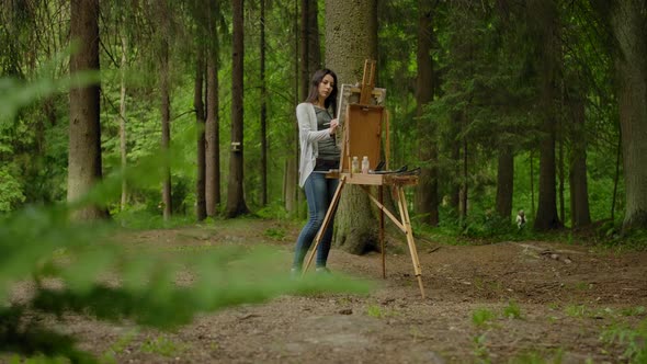 General Wide Shot of a Cute Artist Painting a Landscape on an Easel in the Forest. alt