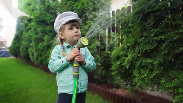 A Small Child is Watering the Plants Near the House with a Hose