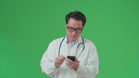 Asian Doctor With Stethoscope Using Mobile Phone While Walking On Green Screen alt