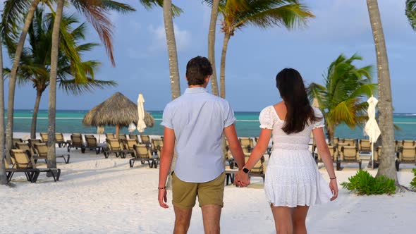 Flirtatious young couple walks on white sandy beach. Magic hour light with tropical palm trees and b alt