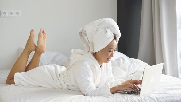 Beautiful Young Chinese Woman Working with a Laptop Lying in Bed at Home Working Remotely Online Due alt