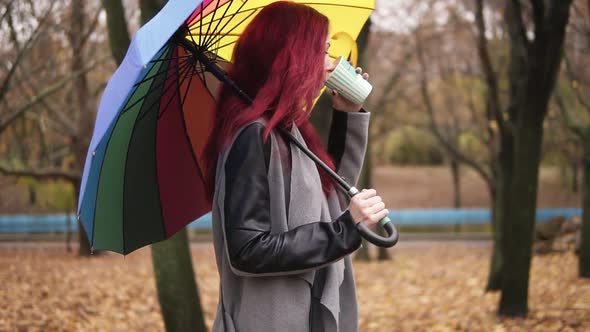 Side View Young Woman with Red Hair Walking in Autumn Park and Drinking Coffee From a Paper Cup alt
