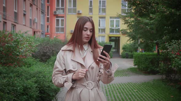 A Shocked Brunette Woman in Beige Coat Looking in Smartphone and Saying Wow and Covering Her Mouth alt