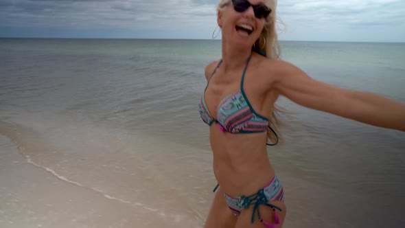 Playful blonde mature woman in sunglasses and bikini running along a beach and looking back at the c, Stock Footage