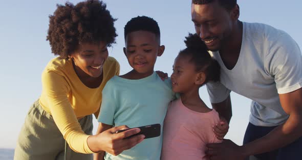 Smiling african american family using smartphone and embracing on sunny beach alt
