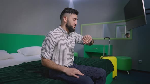 Portrait of Anxious Young Caucasian Man Checking Time on Watch Waiting for Taxi in Hotel Room alt