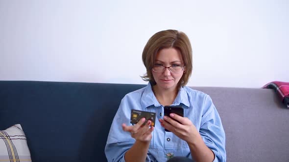 A woman is sitting on the couch in her room and enters data from a bank card into a smartphone
