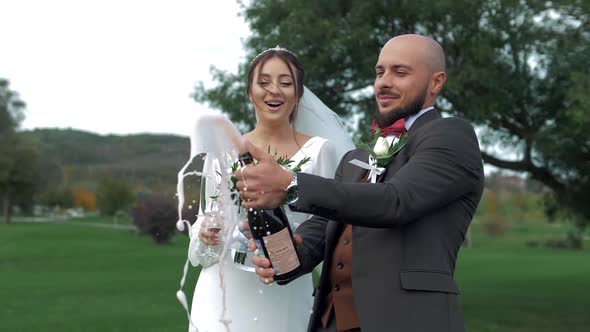 Newlyweds outdoors powerfully opening a bottle of champagne with foam and laughter.