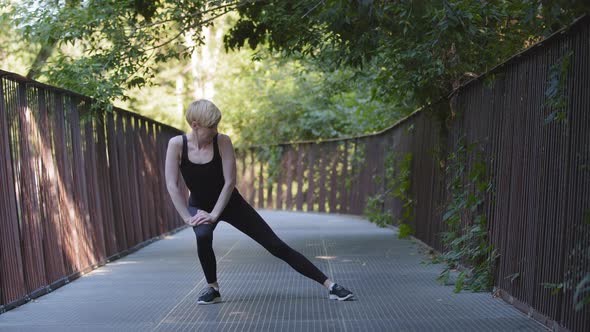 Middleaged Blonde Caucasian Woman Athlete Lady Wears Black Tracksuit Stands on Bridge in City Park alt