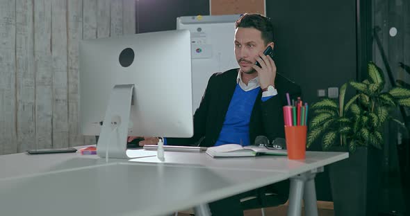 Businessman in the Office in Front of the Computer Talking on the Phone alt