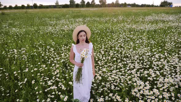 Woman in a White Dress and Hat Walks Through a Field with Daisies in the Evening at Sunset in Summer alt