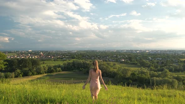 Young woman in pink dress walking on green summer field outdoors. alt
