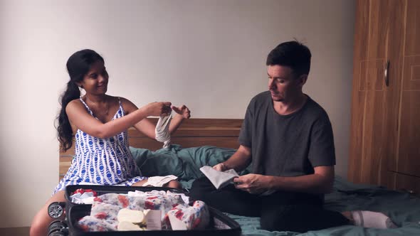 Young Happy Pregnant Couple Sitting on the Bed Packing Things for Baby Preparing Hospital Bag alt